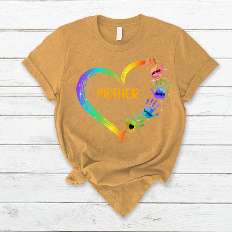 Pu&ograve; includere: Una maglietta color senape con un design a forma di cuore arcobaleno e la parola "MOTHER" all'interno del cuore. Ci sono quattro impronte di mani intorno al cuore, ciascuna con un nome scritto all'interno: Jaidra, Bobby, Theresa e un'impronta di mano pi&ugrave; piccola con un cuore.