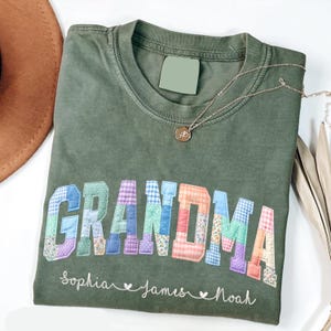 May include: Olive green t-shirt with the word "GRANDMA" in colorful patchwork letters. Below the word are the names "Sophia", "James", and "Noah" in cursive. A gold necklace with a pendant is on the shirt.