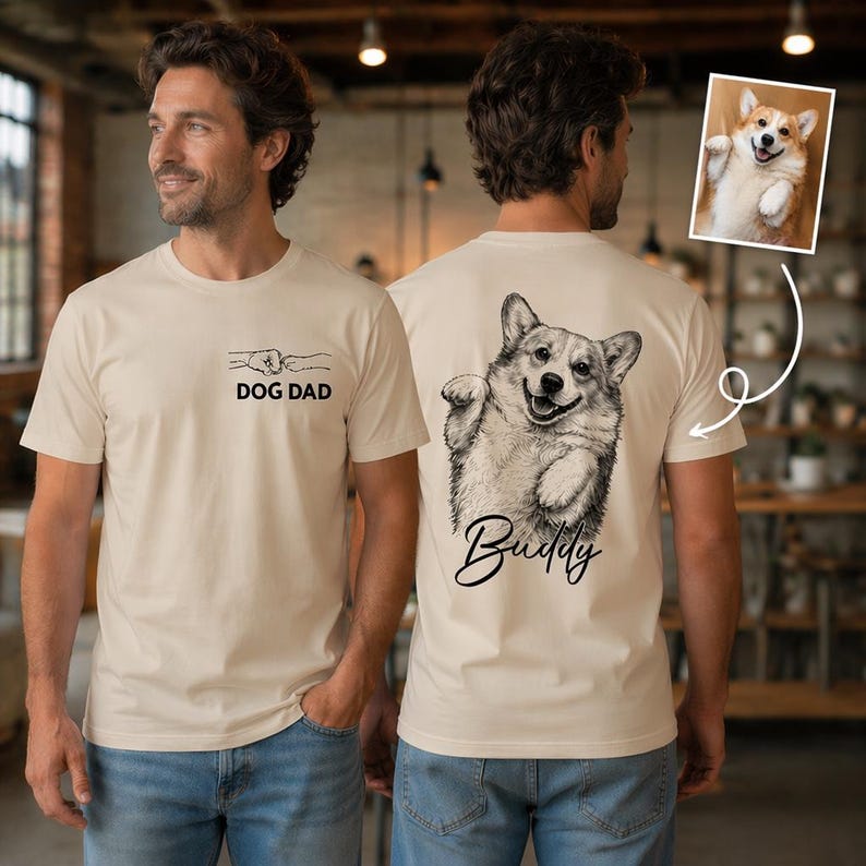 Pode incluir: Uma t-shirt bege de manga curta com um pequeno gr&aacute;fico preto no peito de um punho com o texto DOG DAD. A parte de tr&aacute;s apresenta um grande esbo&ccedil;o a preto e branco de um c&atilde;o Corgi com o nome Buddy escrito em letra cursiva.