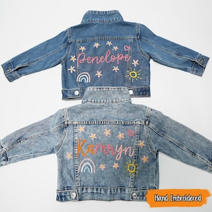 Puede incluir: Dos chaquetas vaqueras con dise&ntilde;os bordados a mano. La chaqueta de arriba tiene el nombre "Penelope" bordado en rosa con un arco&iacute;ris, un sol y estrellas. La chaqueta de abajo tiene el nombre "Kamryn" bordado en naranja con un arco&iacute;ris, un sol y estrellas.