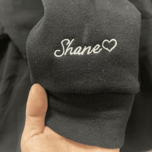 May include: Black sweatshirt with a white embroidered heart and the word "Shane" on the sleeve.