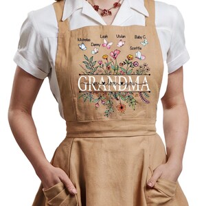 Woman wearing a light brown apron over a white blouse. The apron features the word 'GRANDMA' and a colorful printed design of flowers, leaves, and butterflies. Above the floral illustration are names including Nicholas, Danny, Leah, Vivian, Baby C., and Scottie