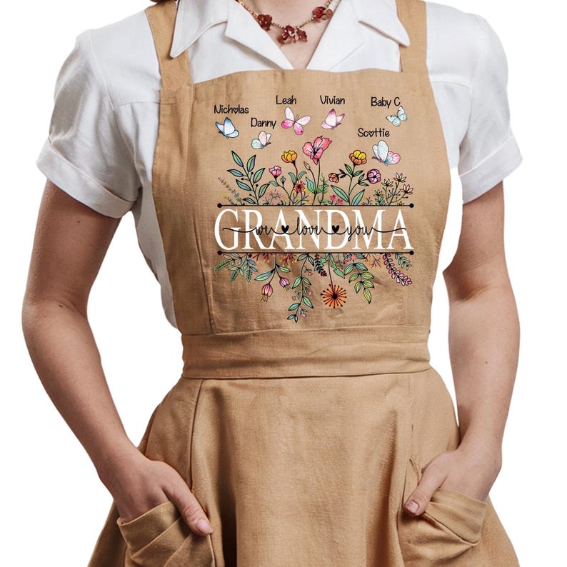 Woman wearing a light brown apron over a white blouse. The apron features the word 'GRANDMA' and a colorful printed design of flowers, leaves, and butterflies. Above the floral illustration are names including Nicholas, Danny, Leah, Vivian, Baby C., and Scottie