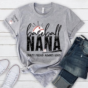 Peut inclure: T-shirt gris avec l'inscription "Baseball Nana" en noir, un graphisme de baseball et la phrase "Crazy Proud Always Loud". Le t-shirt est associ&eacute; &agrave; un jean bleu et des baskets blanches.