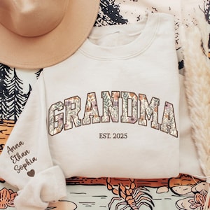 K&ouml;nnte beinhalten: Cremefarbenes Sweatshirt mit dem Wort "GRANDMA" in floralem Design. Der Text "EST. 2025" steht darunter. Der linke &Auml;rmel hat Namen und ein Herz. Ein beiger Hut befindet sich in der oberen linken Ecke. Das Sweatshirt befindet sich auf einer gemusterten Oberfl&auml;che.
