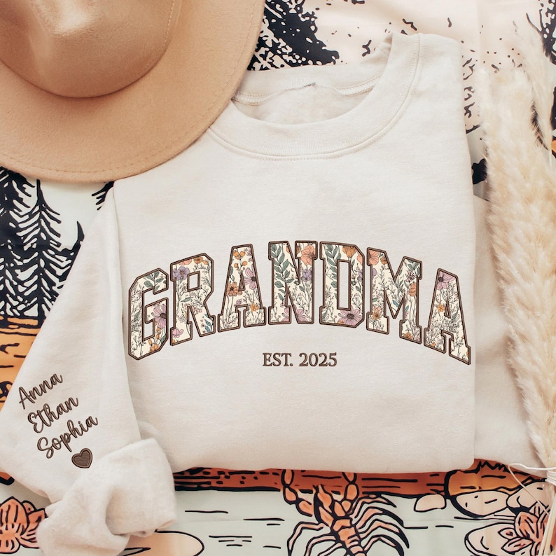 K&ouml;nnte beinhalten: Cremefarbenes Sweatshirt mit dem Wort "GRANDMA" in floralem Design. Der Text "EST. 2025" steht darunter. Der linke &Auml;rmel hat Namen und ein Herz. Ein beiger Hut befindet sich in der oberen linken Ecke. Das Sweatshirt befindet sich auf einer gemusterten Oberfl&auml;che.