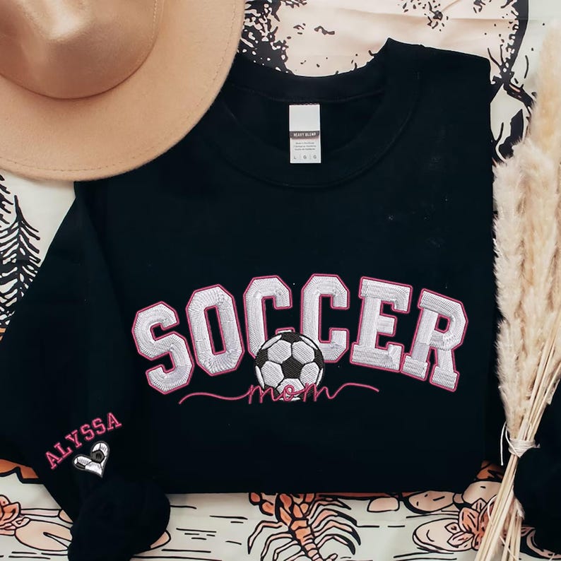 May include: Black sweatshirt with white and pink embroidered text that says "Soccer Mom" with a pink soccer ball in the middle. The left sleeve has the name "Alyssa" embroidered in white with a pink heart.