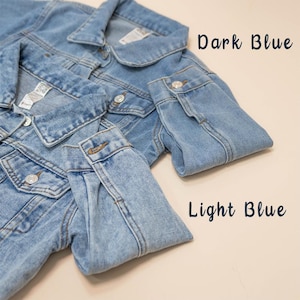 Puede incluir: Dos chaquetas vaqueras, una azul oscuro y otra azul claro, est&aacute;n dispuestas sobre una superficie beige. Las chaquetas est&aacute;n dobladas por la mitad, con las mangas dobladas sobre la parte delantera de las chaquetas.