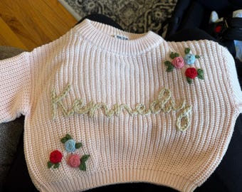 May include: A pink knitted sweater with the word "Kennedy" embroidered in beige yarn. The sweater has floral embroidery with pink, blue, and green accents.