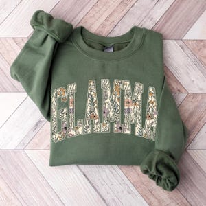 May include: A green sweatshirt with the word "GLAMMA" in a floral design across the chest. The letters are filled with a variety of flowers in shades of cream, orange, and purple. The sweatshirt is folded on a light wood-grain surface.