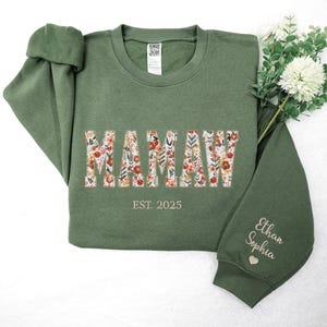 Op de afbeelding: Een groen sweatshirt met het woord "MAMAW" in bloemenprint. De tekst "EST. 2025" staat onder het woord. De mouw heeft de namen "Ethan Sophia" geborduurd in wit met een klein hartje.