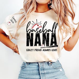 Peut inclure: T-shirt cr&egrave;me avec l'inscription "Baseball Nana". Le design pr&eacute;sente le mot "baseball" au-dessus de "NANA" en gras, avec un effet vieilli. Un ballon de baseball avec un c&oelig;ur est au centre. Le texte "CRAZY PROUD ALWAYS LOUD" est en dessous. Le nom "Emma Jack" est sur la manche.