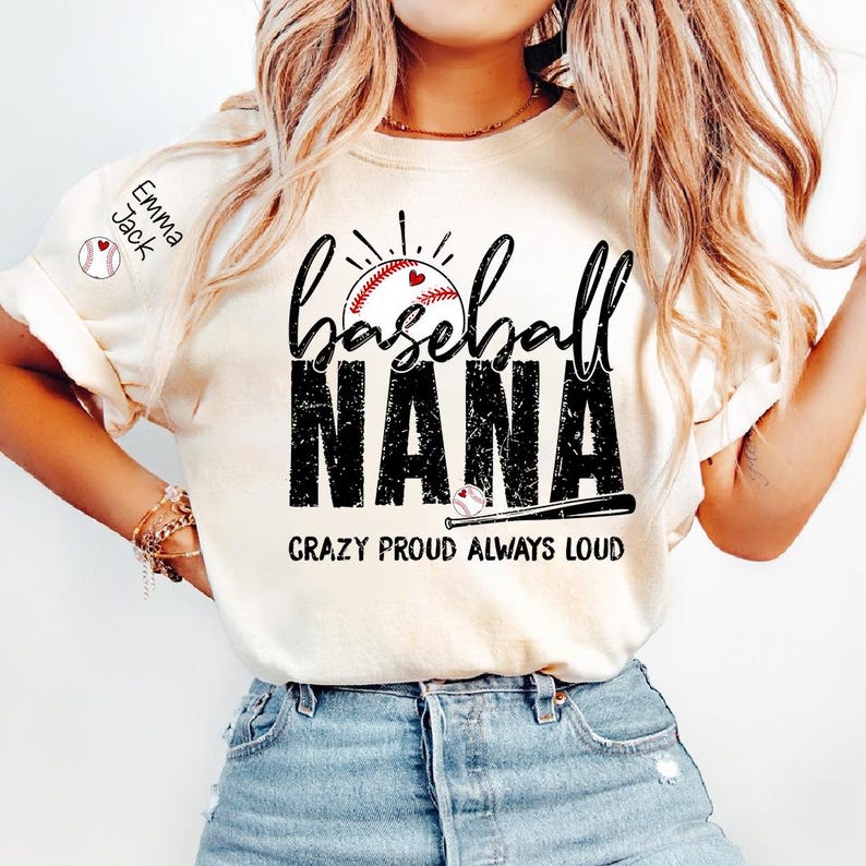 Peut inclure: T-shirt cr&egrave;me avec l'inscription "Baseball Nana". Le design pr&eacute;sente le mot "baseball" au-dessus de "NANA" en gras, avec un effet vieilli. Un ballon de baseball avec un c&oelig;ur est au centre. Le texte "CRAZY PROUD ALWAYS LOUD" est en dessous. Le nom "Emma Jack" est sur la manche.