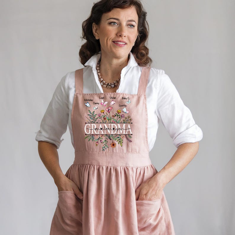 May include: A pink floral apron with the word "Grandma" and the phrase "We love you" printed on it. The apron has pockets on the sides and features a floral design with butterflies and names.