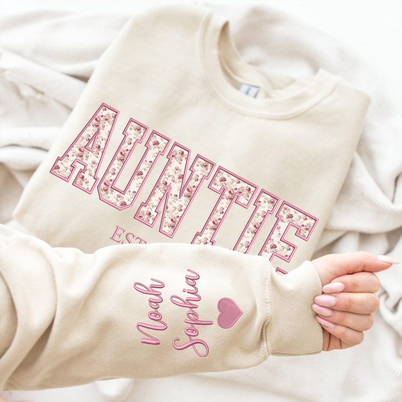 May include: A beige crewneck sweatshirt featuring the word AUNTIE in large block letters with a pink floral pattern and a pink outline. The sleeve displays the names Noah and Sophia in pink script lettering next to a small pink heart icon.