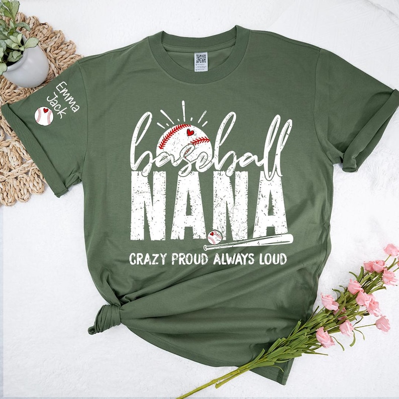Peut inclure: T-shirt vert olive avec l'inscription "Baseball Nana" en blanc. Un graphisme de baseball avec un c&oelig;ur est au-dessus du mot "Nana". Le texte "Crazy Proud Always Loud" est en dessous. La manche porte les noms "Emma Jack" et un graphisme de baseball.