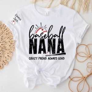 Peut inclure: T-shirt blanc avec un motif sur le th&egrave;me du baseball. Le t-shirt pr&eacute;sente les mots "baseball NANA" dans une police vieillie, un graphisme de baseball et la phrase "CRAZY PROUD ALWAYS LOUD". Le nom "Emma Jack" est imprim&eacute; sur la manche.