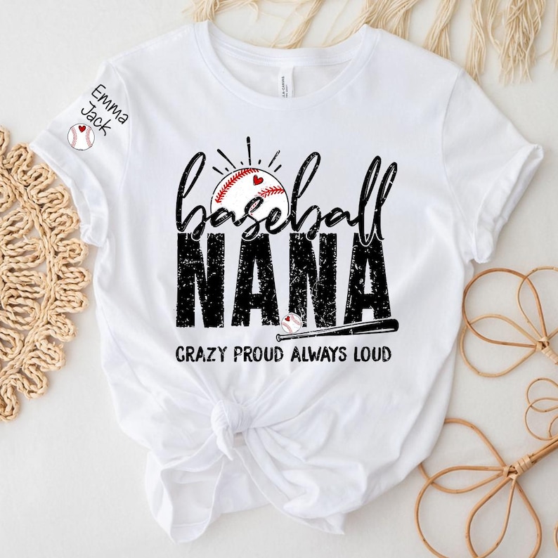 Peut inclure: T-shirt blanc avec un motif sur le th&egrave;me du baseball. Le t-shirt pr&eacute;sente les mots "baseball NANA" dans une police vieillie, un graphisme de baseball et la phrase "CRAZY PROUD ALWAYS LOUD". Le nom "Emma Jack" est imprim&eacute; sur la manche.
