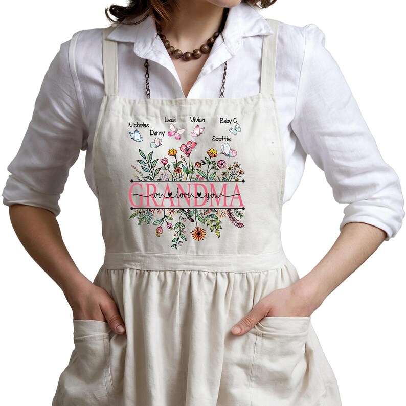 May include: A white apron with a floral design and the text "We love you Grandma" with a pink heart. The apron has pockets on the sides and a variety of butterflies with names printed on them.