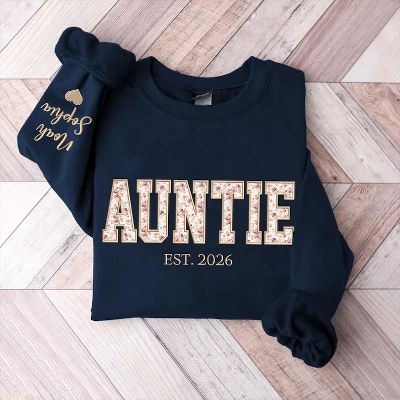 May include: A navy blue crewneck sweatshirt featuring the word AUNTIE in large block letters with a floral pattern fill. Below the word is the text EST. 2026 in gold. The left sleeve displays a gold heart icon above the names Sophia and Noah.