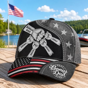 May include: A black and gray baseball cap with a mesh back and a curved brim. The cap features a design with fists and the words "BONUS DAD" and "BEST Bonus Dad EVER". The brim has an American flag design.