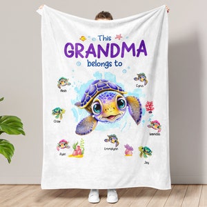 May include: A white fleece blanket with a purple and blue cartoon turtle surrounded by smaller turtles with names. The text "This Grandma belongs to" is printed above the turtle.