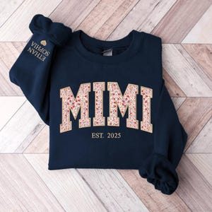 May include: Navy blue sweatshirt with the word "MIMI" in a floral design across the chest. The sleeve has the names "ETHAN SOPHIA" embroidered in gold. The text "EST. 2025" is below the word "MIMI".