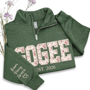 May include: Green quarter-zip sweatshirt with the name "OGEE" in a floral design. The sleeve features the names "Liam, Rory, Grace" and a heart symbol. The text "EST. 2026" is below the name. The sweatshirt is folded.