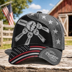 May include: A black baseball cap with an American flag design on the brim. The cap features a graphic of clasped fists with the names "EMMA", "DAVID", "NOAH", and "EMILY". The text "PAWPAW" and "Best Pawpaw Ever" are also visible.