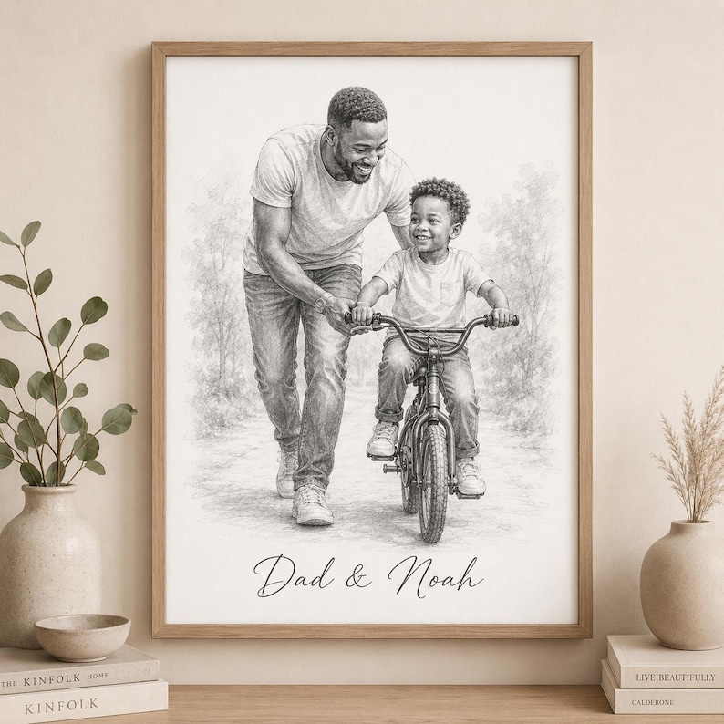 May include: A framed black and white pencil sketch of a father helping his son ride a bicycle. The artwork is signed "Dad & Noah" in cursive script. The frame is a light brown color, and the background is a soft, blurred depiction of trees.