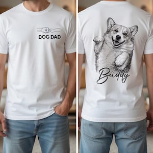 Pode incluir: Uma t-shirt branca com um gr&aacute;fico frontal de um punho humano a bater com uma pata de c&atilde;o e o texto DOG DAD. A parte de tr&aacute;s da t-shirt apresenta um grande esbo&ccedil;o a preto e branco de um c&atilde;o Corgi com o nome Buddy escrito abaixo.