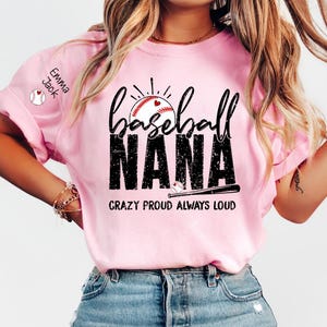 Peut inclure: T-shirt rose clair avec l'inscription "Baseball Nana" en noir vieilli, orn&eacute;e d'un motif de baseball. Le texte "Crazy Proud Always Loud" est imprim&eacute; en dessous. Le texte "Emma Jack" est sur la manche.