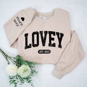 May include: A beige sweatshirt with the word "LOVEY" printed in black with a distressed look. The text "EST. 2023" is printed below the word "LOVEY". The left sleeve has the names "JOHN" and "SOPHIA" printed in black with a heart symbol between the names.