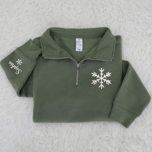 May include: An olive green quarter-zip sweatshirt with a white snowflake design on the front. The left sleeve has the name "Sophia" embroidered in white, along with a small snowflake. The sweatshirt is folded on a white, fluffy surface.
