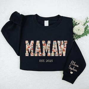 Op de afbeelding: Zwart sweatshirt met het woord "MAMAW" in bloemenprint letters. De tekst "EST. 2025" staat onder het woord. De mouw heeft de namen "Ethan Sophia" geborduurd in gouden draad.