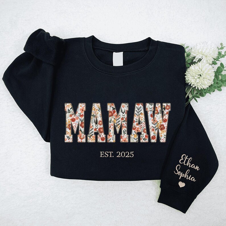 Op de afbeelding: Zwart sweatshirt met het woord "MAMAW" in bloemenprint letters. De tekst "EST. 2025" staat onder het woord. De mouw heeft de namen "Ethan Sophia" geborduurd in gouden draad.