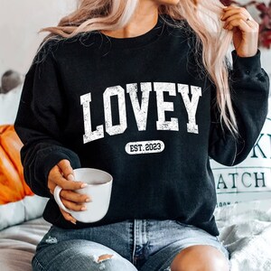 May include: Black sweatshirt with white text that reads "LOVEY EST. 2023".