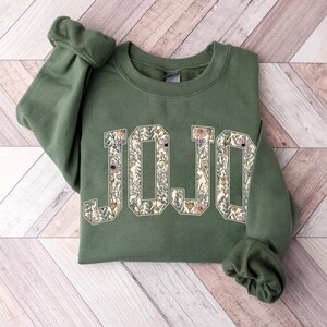 May include: Green sweatshirt with the name "JOJO" in large, floral-filled letters. The letters are outlined in a darker green, with a cream-colored background. The floral design includes purple, orange, and cream-colored flowers.