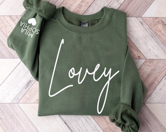 May include: A green sweatshirt with the word "Lovey" written in white cursive on the front. The sweatshirt has a crew neck and long sleeves.