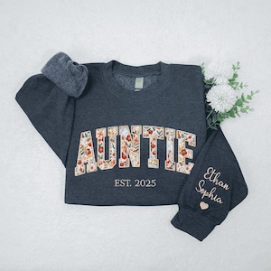 May include: A dark grey sweatshirt with the word "AUNTIE" in floral print. The text "EST. 2025" is below the word "AUNTIE". The left sleeve has the names "Ethan Sophia" and a small heart.