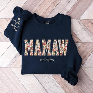 Op de afbeelding: Navyblauwe trui met een bloemenprintontwerp met de tekst "MAMAW EST. 2025".