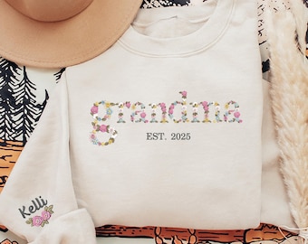 Könnte beinhalten: Ein hellbeiger Pullover mit dem Wort "grandma", das in bunten Blumen geschrieben ist. Der Text "EST. 2025" ist unter dem Wort "grandma" gedruckt. Der Pullover hat ein kleines gesticktes Blumenmuster auf dem linken Ärmel mit dem Wort "Kelli" darüber.