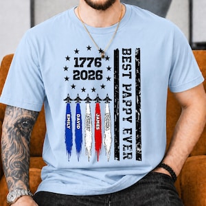 May include: A light blue t-shirt featuring a graphic with the text 1776 2026, five fighter jets with colored smoke trails, and the text BEST PAPPY EVER. The design includes a circle of stars and vertical stripes in a distressed black print.
