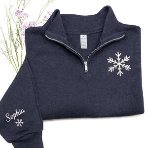 May include: A navy blue quarter-zip sweatshirt with a white snowflake embroidered on the chest. The left sleeve has the name "Sophia" embroidered in white, along with a small snowflake. The sweatshirt has a silver zipper.