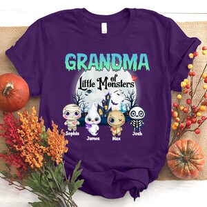 K&ouml;nnte beinhalten: Lilafarbenes T-Shirt mit dem Text "Grandma of Little Monsters" in einem wei&szlig;en Kreis mit Mond und Sternen. Unter dem Kreis befinden sich vier Comic-Monster mit den Namen Sophia, James, Max und Josh.