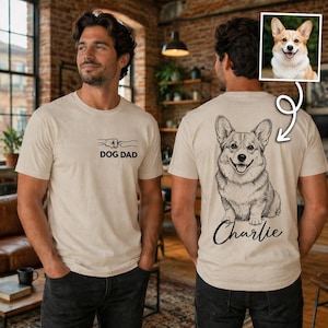 Pode incluir: Uma t-shirt bege com um pequeno gr&aacute;fico no peito de um punho humano a bater com uma pata de c&atilde;o e o texto DOG DAD. A parte de tr&aacute;s da t-shirt apresenta uma grande ilustra&ccedil;&atilde;o de linhas de um c&atilde;o Corgi com o nome Charlie escrito por baixo.
