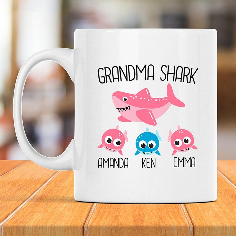 May include: White ceramic mug with a pink shark illustration and the text "Grandma Shark". Below the shark are three smaller pink and blue shark illustrations with the names "Amanda", "Ken", and "Emma" underneath.