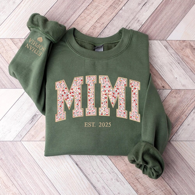 May include: Green sweatshirt with the word "MIMI" in a floral pattern across the chest. The text "EST. 2025" is below the name. The left sleeve has the names "ETHAN SOPHIA" embroidered in gold.