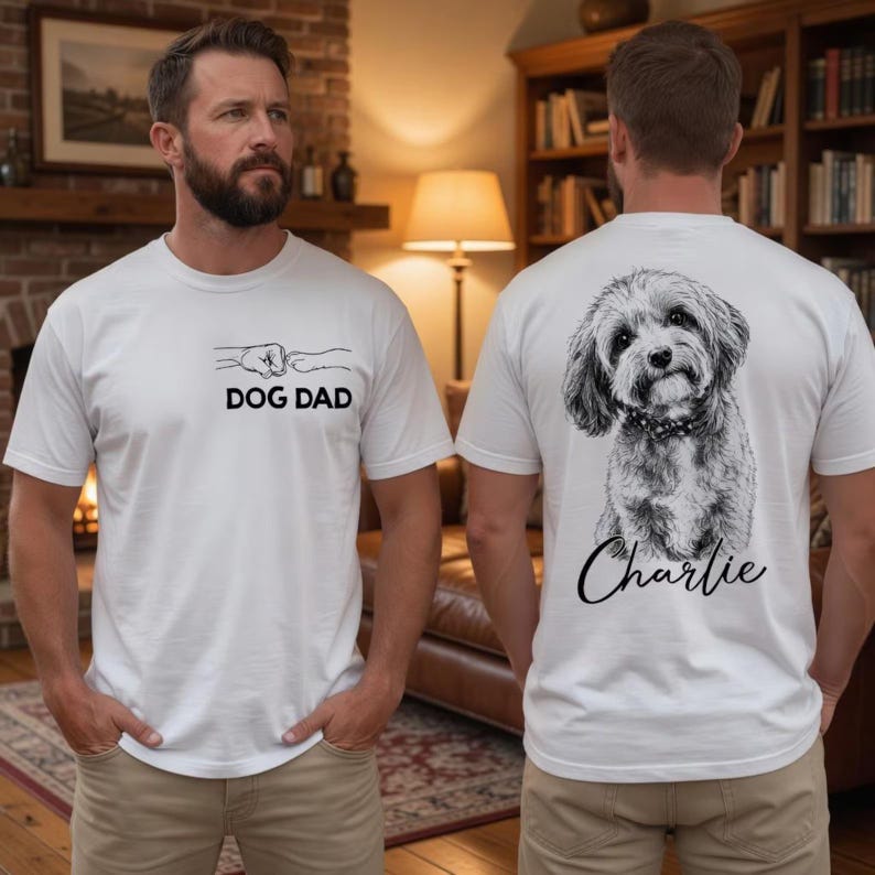 Pode incluir: Uma t-shirt branca de manga curta com um gr&aacute;fico preto na frente de um punho humano a bater numa pata de c&atilde;o com o texto DOG DAD. As costas apresentam um esbo&ccedil;o detalhado a preto e branco de um c&atilde;o com o nome Charlie impresso abaixo.