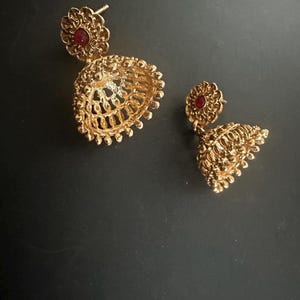 Op de afbeelding: Goudkleurige filigraan oorbellen met een bloemmotief. Elke oorbel heeft een kleine, ronde, rode edelsteen aan de bovenkant en een klokvormig, opengewerkt ontwerp eronder. De oorbellen worden op een donkere ondergrond getoond.